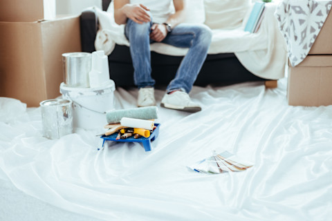 A man sits on a couch with cardboard boxes sitting on either side. A sheet is laid down in front of the couch, with painting tools and samples laying on it.