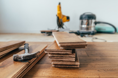 In a room that’s being refloored, a closeup of a pile of oak floor panels sitting on the part of the floor that’s been done. A hammer is sitting next to the pile and in the background we can see equipment.