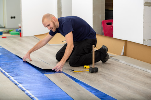 A worker installs click-lock LVP flooring on a renovation site. A few work tools lie next to him.