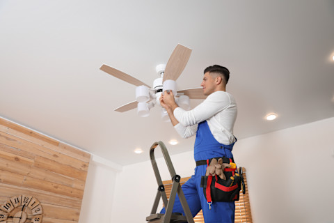 A pro on a ladder with a toolbelt installs a ceiling fan that has lights attached.