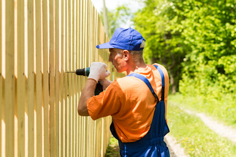 Side angle of a worker using a power drill to install a wooden fence.