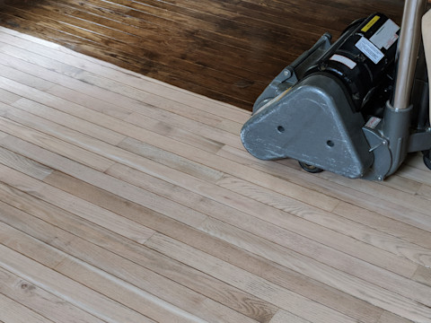 A worker kneeling on a hardwood floor pours wood filler from a bottle onto a damaged area of the floor during a refinishing project.