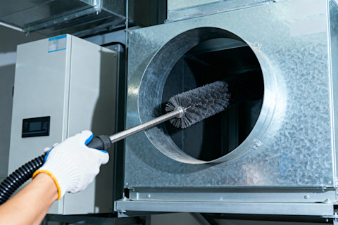 We can see a worker’s gloved hands holding a duct cleaning brush attached to a flexible rod/drill extension, which they are inserting into a large round metal air duct opening.