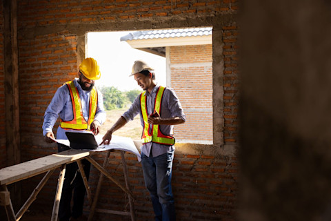 Two workers wearing collared shirts, work vests and hard hats look over architectural plans on a construction site in a room with an old brick wall.
