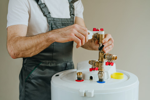 A water heater installer wearing overalls wraps plumbing tape around a fitting on a water heater.