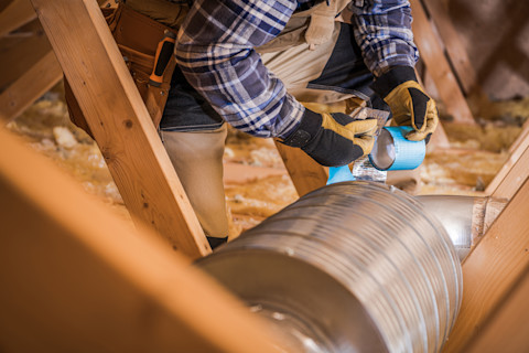 A worker is bent over attic ductwork, tearing off a piece of tape from a roll. The attic’s wooden beams and insulation are visible.