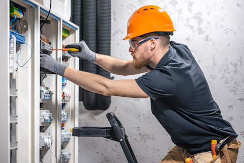 A male electrician works on a switchboard with fuses. He inspects electrical connections inside a control panel.