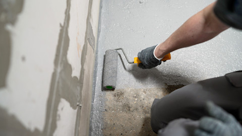 A worker waterproofs a cement basement floor using a paint roller. Downward shot where only the worker’s hand and knee are visible.