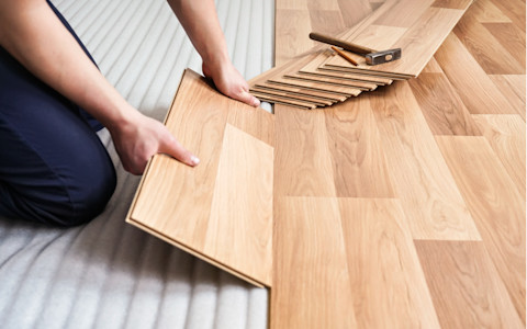 This image shows a close-up of a worker installing laminated flooring on top of underlay, carefully aligning and securing the planks for a seamless finish. There is a hammer and pencil on top of one of the planks that is yet to be installed.
