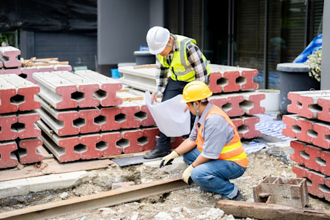 Two workers in hard hats and reflective vests compare groundwork to architectural plan.