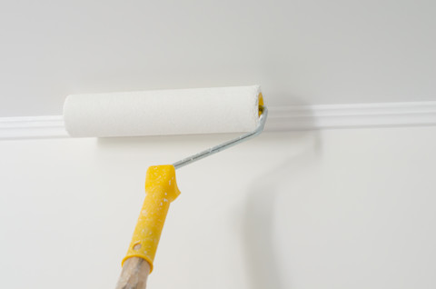 Closeup of a paint roller with a yellow handle applying white paint or primer to a ceiling.