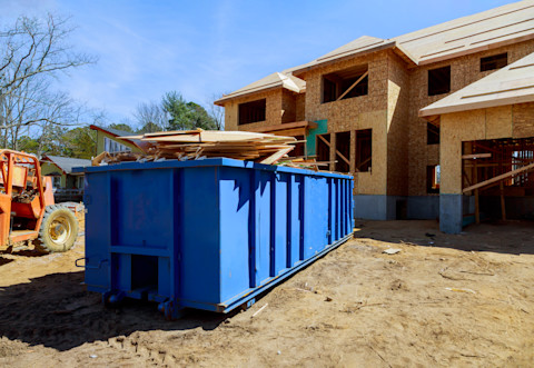This image shows a blue dumpster container in the front yard of a house that is undergoing construction. The container is filled with scrap materials from the build.