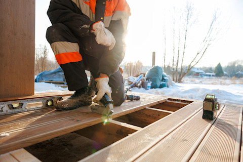 A worker in protective clothing kneels to work on a wooden deck using a level and other tools. Snow is visible in the background.