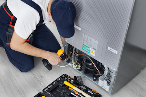 A repair specialist wearing a navy blue cap and overalls is crouched behind a fridge and uses a drill for repairs.