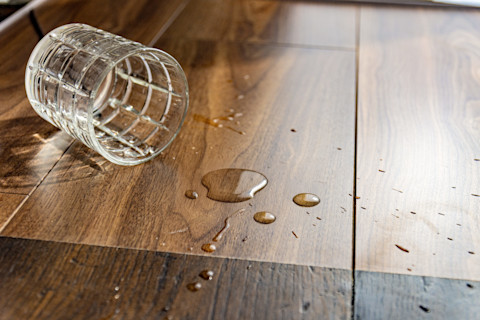 A glass of water tipped on its side next to some drops of water on a wooden laminate floor.
