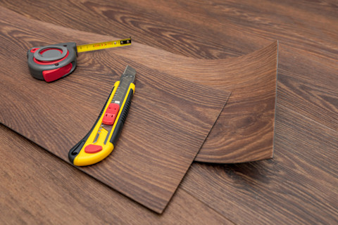 Some vinyl floor panels lay on some installed floor panels of the same design, with a measuring tape and box cutter on top.