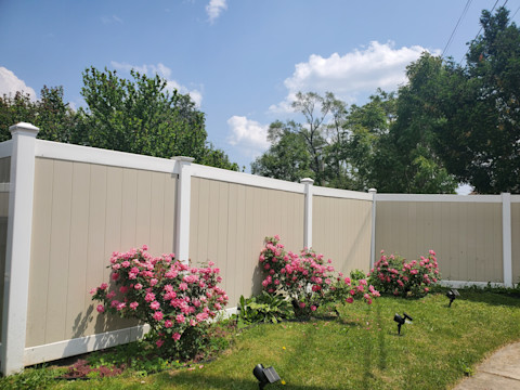 A beige with white solid fence in a garden with pink flower bushes and grass.