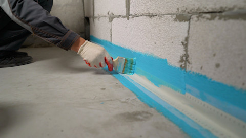 Closeup of a worker’s gloved hands waterproofing the bottom of a concrete wall with blue sealant.