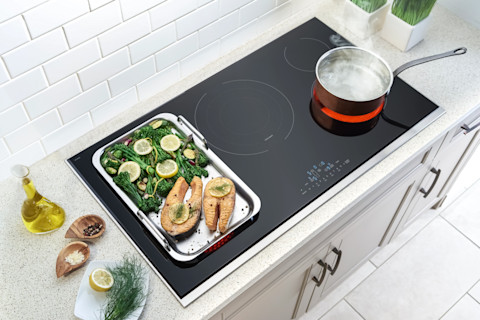 Aerial shot of an electric stovetop with a tray of cooked salmon and greens and a boiling pot of water. Some herbs, seasoning and oil are on the counter to the left of the stove.
