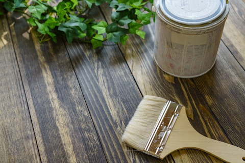 Closeup of a paintbrush and can of paint sitting on a treated wooden surface. Ivy is visible along the upper edge of the photo.