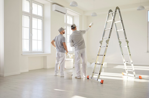 Two professional painters in conversation while looking at a wall.They’re in an all-white room, and a ladder and a few buckets are visible.
