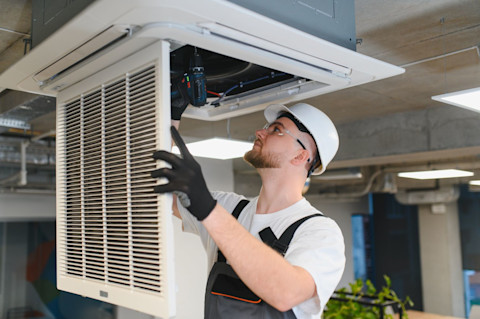 An air con installer wearing overalls, hard hat, safety glasses and gloves performs work on a ceiling AC unit using a powerdrill.
