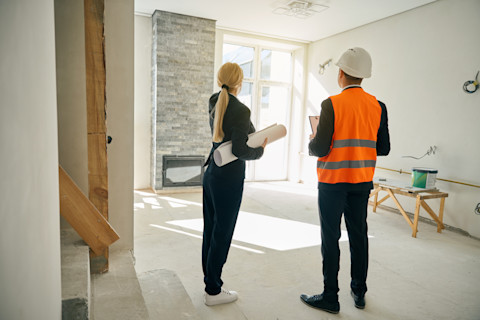 A woman holds a piece of rolled up posterboard under her arm and stands next to a construction worker holding a clipboard in a room under renovation.