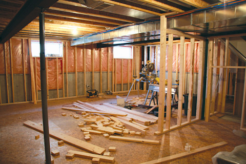 A basement mid-renovation with insulation visible, with a saw set up on a workbench in the centre of the space. Lumber lies on the floor in front of the workbench.