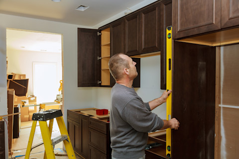 This image shows a cabinet maker or carpenter using a tape measure to help him install dark wood cabinets in a kitchen.