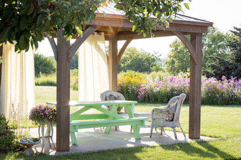 This image shows a wooden gazebo in a backyard. The lighting is soft and the grass is green and pink flowers are in bloom, suggesting that the photo has been taken in the summertime. There is a light green picnic table beneath the gazebo and wicker chairs.