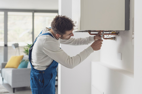 A man in blue overalls is fixing the pipes of a wall-mounted boiler in a bright, modern living room. The room has a gray sofa with colourful cushions and large windows.