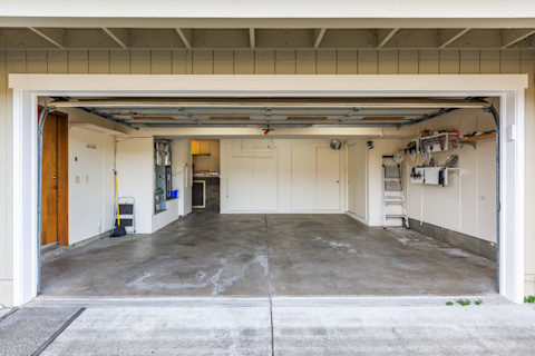 Empty two-car garage with an open door, showing a bare, stained concrete floor, a stepladder and ladder, a broom, and a wall-mounted tool shelf.
