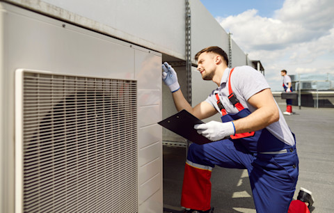 A pro holding a clipboard kneels as he examines an HVAC unit on a roof.