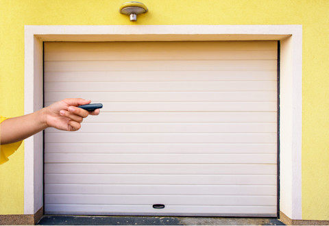 Frontal shot of a white garage door in a yellow house, with a hand holding a remote coming in from the left side of the frame.