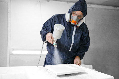 A pro in full protective gear uses a spray gun to apply white paint to a kitchen cabinet door.