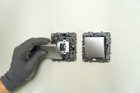 Closeup of an electrician’s gloved hands installing a light switch. One is already installed, but the faceplates are missing.