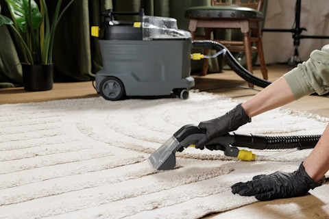 A gloved worker uses an extraction machine to deep clean a beige rug in a living room.