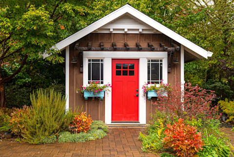 Frontal shot of a dark brown garden shed with a bright wooden door and flower boxes on the windows. Set in a landscaped garden with red and green bushes, with trees in the back.