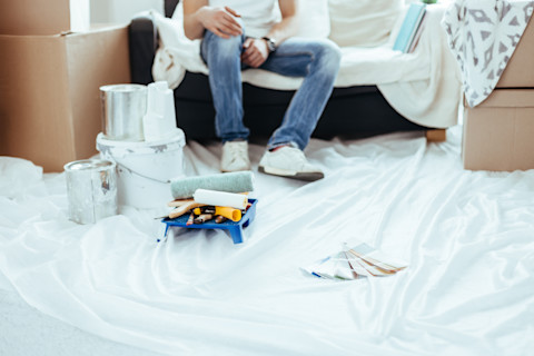 A man sits on a couch with cardboard boxes sitting on either side. A sheet is laid down in front of the couch, with painting tools and samples laying on it.