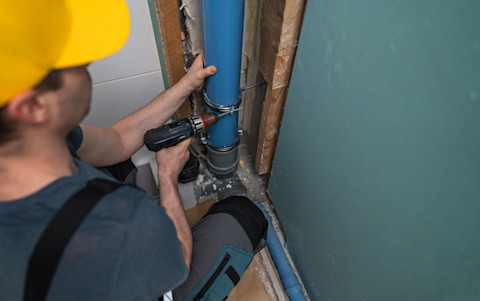 A worker crouches in what appears to be the corner of a basement, holding onto a drain pipe system with one hand and a power drill with the other.