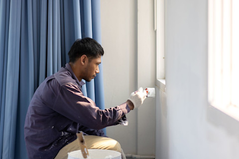 A painter crouches to paint under a window in the corner of a room with white walls. A blue curtain is visible behind him.