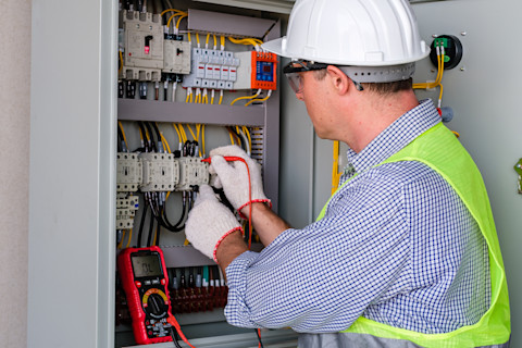 This image shows an electrician installing a new electrical control panel. He is wearing safety gloves, a high visibility vest, a hard hat and safety glasses.