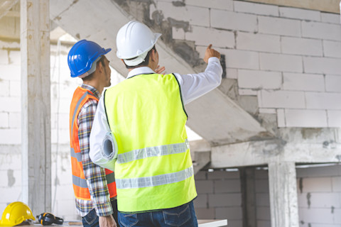 This image shows a general contractor and a builder wearing hard hats and high visibility vests in a building that is being renovated. They are pointing to something out of shot and discussing renovation plans.