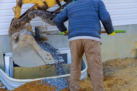A worker uses a shovel to push gravel from a bucket loader into the space in front of an egress window.