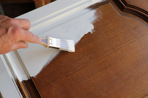 Closeup of a brown wooden kitchen cabinet being painted white.